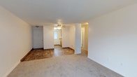 This single-floor residence offers a practical and open layout designed for comfortable everyday living. The main living area is finished with plush carpet flooring, creating a warm and inviting atmosphere, while clean white walls and a textured ceiling contribute to a bright, neutral aesthetic throughout.

A defining architectural feature of the space is a structural column that subtly divides the living area from the adjacent dining and kitchen zone, maintaining an open feel while providing a natural sense of separation between spaces. The kitchen area transitions to wood-look hard flooring and features warm, honey-toned cabinetry that adds a traditional character to the space. A window in the kitchen allows natural light to filter in, brightening the area throughout the day.

A ceiling fan with a light fixture is centrally positioned overhead, providing both air circulation and ambient lighting. The entry door is visible from the living area, offering a straightforward and accessible layout. A hallway leads further into the residence, hinting at additional rooms beyond. The combination of carpet and hard flooring, along with the open sightlines, makes this a functional and well-organized space suited for a range of living arrangements.
