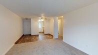 This single-floor residence offers a practical and open layout designed for comfortable everyday living. The main living area is finished with plush carpet flooring, creating a warm and inviting atmosphere, while clean white walls and a textured ceiling contribute to a bright, neutral aesthetic throughout.

A defining architectural feature of the space is a structural column that subtly divides the living area from the adjacent dining and kitchen zone, maintaining an open feel while providing a natural sense of separation between spaces. The kitchen area transitions to wood-look hard flooring and features warm, honey-toned cabinetry that adds a traditional character to the space. A window in the kitchen allows natural light to filter in, brightening the area throughout the day.

A ceiling fan with a light fixture is centrally positioned overhead, providing both air circulation and ambient lighting. The entry door is visible from the living area, offering a straightforward and accessible layout. A hallway leads further into the residence, hinting at additional rooms beyond. The combination of carpet and hard flooring, along with the open sightlines, makes this a functional and well-organized space suited for a range of living arrangements.