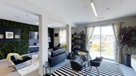 This single-floor space showcases a contemporary open-concept design that seamlessly integrates the living and dining areas into a cohesive, visually striking environment. The interior is defined by a bold monochromatic palette accented with vibrant yellow touches, creating a dynamic yet balanced aesthetic throughout.

The living area features a plush dark sofa adorned with yellow accent pillows, set against a striking living green wall that adds a natural, textural element to the space. A flat-screen television is mounted nearby, completing a stylish and functional lounge setting. The room benefits from ample natural light streaming through multiple windows dressed with flowing white curtains.

The dining area is anchored by a round glass-top table surrounded by modern black chairs with natural wood legs, all positioned atop a bold black-and-white striped area rug that adds graphic energy to the floor plane. A sleek open shelving unit provides both storage and display space, adding depth to the room.

Linear ceiling lighting fixtures run overhead, providing clean, modern illumination that complements the overall design language. A large sliding glass door opens directly onto an outdoor terrace furnished with seating, offering an inviting extension of the interior living space with open views of the surrounding area.