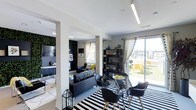 This single-floor space showcases a contemporary open-concept design that seamlessly integrates the living and dining areas into a cohesive, visually striking environment. The interior is defined by a bold monochromatic palette accented with vibrant yellow touches, creating a dynamic yet balanced aesthetic throughout.

The living area features a plush dark sofa adorned with yellow accent pillows, set against a striking living green wall that adds a natural, textural element to the space. A flat-screen television is mounted nearby, completing a stylish and functional lounge setting. The room benefits from ample natural light streaming through multiple windows dressed with flowing white curtains.

The dining area is anchored by a round glass-top table surrounded by modern black chairs with natural wood legs, all positioned atop a bold black-and-white striped area rug that adds graphic energy to the floor plane. A sleek open shelving unit provides both storage and display space, adding depth to the room.

Linear ceiling lighting fixtures run overhead, providing clean, modern illumination that complements the overall design language. A large sliding glass door opens directly onto an outdoor terrace furnished with seating, offering an inviting extension of the interior living space with open views of the surrounding area.