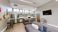 This contemporary single-floor space showcases a thoughtfully designed open-plan layout that seamlessly blends the kitchen and living areas into one cohesive, functional environment. The kitchen is a standout feature, boasting sleek wood-grain lower cabinetry paired with crisp white quartz countertops and a full subway tile backsplash. Open walnut floating shelves provide both display and storage space, while stainless steel appliances — including a range, refrigerator, and dishwasher — lend a polished, professional feel. A row of clerestory windows runs the length of the kitchen wall, flooding the space with natural light. A freestanding industrial-style island with a metal frame and reclaimed wood lower shelf anchors the center of the room. The living area features light hardwood flooring that flows throughout, soaring ceilings, and a wall-mounted flat-screen TV. A high-top bistro table with bar stools provides a casual dining option. The most dramatic architectural element is the floor-to-ceiling glass curtain wall at the entry, framing views of lush greenery outside and creating a bright, welcoming atmosphere. The overall design is modern and minimalist, with a warm, inviting character throughout.