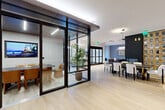 Located at 1350 Dresden Dr NE in Brookhaven, Georgia, this thoughtfully designed single-floor amenity space offers a refined and functional environment for gathering, working, and relaxing. Light hardwood flooring flows throughout, lending warmth and continuity to the open layout.

A glass-walled conference room with dark steel framing anchors one side of the space, providing a semi-private setting equipped with a large display screen and seating for multiple guests. A dramatic double-sided fireplace with a sleek dark surround and white hearth platform serves as a striking architectural centerpiece, visually dividing the lounge and dining areas while adding an inviting ambiance.

The lounge area is outfitted with stylish seating and a mounted television, while a separate dining zone features a dark communal table surrounded by upholstered chairs beneath modern pendant lighting. A floor-to-ceiling display wall with warm wood cabinetry adds a distinctive design element to the dining area.

A wet bar nook showcases deep navy cabinetry, open brass shelving, and bold dark tile backsplash — ideal for entertaining. Large windows frame views of a landscaped courtyard complete with a pergola and lush plantings, filling the space with natural light and a connection to the outdoors.