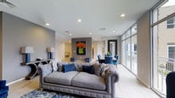 Located at 4328 Ridge Avenue in the Roxborough neighborhood of Philadelphia, PA, this single-floor space offers a thoughtfully designed open-concept layout that seamlessly blends living and dining areas. The interior is characterized by clean, contemporary finishes, including smooth light-toned flooring and recessed lighting throughout, creating a bright and welcoming ambiance.

The living area is anchored by a tufted Chesterfield-style sofa in a neutral gray tone, complemented by navy blue accent ottomans and coordinated throw pillows that add depth and color to the space. A bold piece of wall art featuring vibrant orange florals serves as a striking focal point against the accent wall, adding a touch of personality to the otherwise refined palette.

One of the standout features of this space is the expansive floor-to-ceiling window wall, which floods the interior with abundant natural light and provides an open, airy feel. The dining area transitions naturally from the living space, furnished with a sleek table and blue upholstered chairs beneath a pendant light fixture.

The overall design aesthetic is modern and cohesive, with a carefully curated color scheme of grays, blues, and warm neutrals that lend the space a polished yet inviting character.