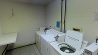 This single-floor laundry room provides a practical and functional space designed for everyday laundry needs. The room features white tile flooring and neutral-toned walls, creating a clean and utilitarian environment. A top-loading Kenmore washing machine is prominently positioned in the foreground, paired with a set of front-accessible dryer units along the adjacent wall. The machines are arranged to allow reasonable ease of movement within the space. A notice board with informational signage is mounted on the wall, likely displaying usage guidelines or building rules. Vertical piping runs along one wall, indicative of the room's utility infrastructure. Baseboard heating is also present, ensuring the space remains functional year-round. The overhead lighting provides adequate illumination throughout the room. While compact in scale, the layout is straightforward and efficient, making it well-suited for its intended purpose. The overall ambiance is no-frills and practical, reflecting a shared or building-use laundry facility focused on functionality over aesthetics.