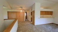 This single-floor space is designed with an open, flowing layout that maximizes both comfort and functionality. The living area is anchored by vaulted ceilings that lend a sense of spaciousness and airiness to the interior. A ceiling fan with a light fixture serves as a practical centerpiece overhead, providing both circulation and ambient lighting.

A defining architectural feature is the partial wall with a built-in pass-through counter that connects the living area to the kitchen, creating a sense of openness while maintaining a defined cooking space. The kitchen is equipped with warm honey-toned wood cabinetry that extends to the upper walls, offering generous storage. A white refrigerator is visible within the kitchen, which continues the neutral, clean aesthetic throughout.

Plush wall-to-wall carpeting in a warm beige tone flows through the main living areas, contributing to the cozy and inviting ambiance of the space. The hallway leading toward the rear of the unit provides access to additional rooms and features sliding wood-panel closet doors, adding to the cohesive warm wood accents found throughout. Neutral white walls complement the natural tones of the cabinetry and flooring, creating a bright, welcoming atmosphere ideal for everyday living.
