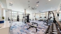 Located at 1840 SW 43rd St in Gainesville, FL, this fitness center is a thoughtfully designed, single-story facility divided into two distinct exercise rooms that together offer a comprehensive workout experience.

The main exercise room is a spacious, open area featuring a geometric blue-and-gray patterned carpet that adds a modern aesthetic. Large windows line the perimeter walls, flooding the space with natural light and offering views of the surrounding greenery. Oversized ceiling fans circulate air throughout. The room is outfitted with a full cable machine, a power rack with a bench press, a complete dumbbell rack, multiple treadmills, elliptical cross-trainers, and adjustable benches. Wall-mounted flat-screen TVs and floor-to-ceiling mirrors enhance both functionality and the sense of space.

The second exercise room offers a more intimate environment designed for functional and cardio training. It features warm hardwood-style flooring, a large wall mirror, and a wall-mounted flat-screen TV. Equipment includes a spin bike, battle ropes, medicine balls, a heavy bag, and resistance accessories stored on a dedicated rack. A large ceiling fan and recessed lighting keep the room bright and comfortable. White panel doors connect the two rooms, creating a seamless and well-organized fitness facility overall.