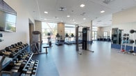 This contemporary fitness center is thoughtfully designed to support a full range of workout routines across multiple functional zones. The main exercise floor features a generous open layout with light-toned flooring, a drop ceiling with recessed and panel lighting, and neutral walls accented by a distinctive navy blue base trim that runs throughout the space. Floor-to-ceiling windows along one wall flood the cardio area with natural light and offer open outdoor views, creating an energizing atmosphere for exercise.

The cardio zone is lined with a row of modern treadmills positioned directly in front of the large windows, providing users with bright, scenic workout conditions. Nearby, cable and functional training machines are strategically placed within the open floor plan, complemented by a dumbbell rack and flat benches for free weight training. Full-length mirrors are mounted along key walls throughout the facility, enhancing the sense of space and allowing users to monitor their form.

A dedicated cycling studio is separated from the main floor and features a smooth blue rubberized floor, a wall of mirrors, and a line of stationary spin bikes. The studio also benefits from natural light through glass doors that open to the exterior. A wall-mounted television is present in the transitional area between zones, and the overall layout ensures clear sightlines and easy movement throughout the facility.