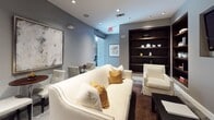 This thoughtfully designed single-floor space combines modern aesthetics with a warm, inviting atmosphere. The entry corridor sets an immediately professional tone, featuring terrazzo flooring, glass-panel doors with push-to-open accessibility hardware, and recessed lighting that guides visitors seamlessly into the interior. The hallway's clean lines and neutral gray palette establish a cohesive design language carried throughout the space.

At the heart of the layout is a well-appointed lounge area that balances elegance with comfort. A generously sized white upholstered sofa with mustard and neutral accent pillows serves as the focal point, complemented by a matching white armchair and a sleek dark coffee table. Underfoot, a rich brown area rug adds warmth and texture against the neutral flooring. Recessed ceiling lights provide even, ambient illumination throughout.

Built-in dark wood shelving units are a standout architectural feature, displaying decorative objects, books, and accent pieces in an organized yet curated arrangement. A wall-mounted flat-screen television is integrated seamlessly into the design alongside the shelving. Framed artwork on the walls adds a refined cultural touch. A small café-style seating area with round tables and chairs is visible in the background, offering an additional functional zone within this versatile and well-considered space.