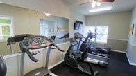 This single-level fitness center offers a focused workout environment designed to accommodate essential cardio training. The space is outfitted with a selection of cardio equipment, including a treadmill, a stationary upright bike, and an elliptical machine, providing residents or members with a variety of exercise options. A large wall mirror spans one side of the room, visually expanding the space while allowing users to monitor their form during workouts. A wall-mounted flat-screen television is positioned for easy viewing from the equipment, adding an entertainment element to exercise sessions. Natural light filters into the space through a window overlooking outdoor greenery, creating a bright and pleasant atmosphere. The ceiling features a fan with a light fixture, contributing to ventilation and comfort during workouts. Dark flooring provides a practical, durable surface suited for fitness use. The light-toned walls and overall layout keep the space feeling open and functional, making it a convenient and well-appointed facility for daily fitness routines.