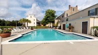 Located at 17931 GA Highway 67 S in Statesboro, Georgia, this outdoor amenity area centers around a large, rectangular swimming pool with clear blue water, surrounded by an expansive concrete sun deck. The pool area is generously proportioned, offering ample space for both swimming and relaxation. Numerous lounge chairs and chaise lounges are arranged along the deck, providing plenty of seating for sunbathing and outdoor leisure.

The space is enclosed by a sturdy black iron fence and gate, offering both security and a polished aesthetic. Lush hedgerows and potted palm trees add a touch of greenery and tropical character to the environment. The surrounding multi-story buildings feature neutral stucco exteriors with traditional shuttered windows, lending the property a classic Southern architectural style.

The pool area opens to views of mature trees and natural Georgia landscape beyond the perimeter fence, creating a pleasant backdrop. The grounds are well-maintained, with manicured landscaping including flowering plants and ivy-covered walls along the walkways. The overall ambiance is that of a comfortable, resort-style outdoor retreat well-suited for warm Southern summers, offering residents a welcoming space to relax and enjoy the outdoors.
