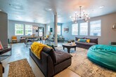 This single-floor communal space is designed to accommodate a variety of social activities within an open, well-connected layout. The main lounge area is furnished with plush, dark upholstered sofas accented by bold yellow and teal throw pillows, anchored by patterned area rugs over polished concrete flooring. A structural column divides the space subtly without disrupting its open feel. Ambient and task lighting is provided by a striking multi-bulb sputnik-style chandelier, recessed ceiling lights, and wall-mounted sconces.

One end of the space transitions into a media lounge, featuring a wall-mounted flat-screen television set against a warm mustard-yellow accent wall, complemented by bean bag seating and additional sofa arrangements. Natural light enters through multiple windows fitted with horizontal blinds throughout the space.

A game and dining area sits toward the rear, furnished with high-top bar tables, industrial-style stools, and a pool table beneath pendant lighting. The kitchenette nearby includes dark espresso cabinetry, a subway tile backsplash, white countertops, a stainless steel sink, microwave, and refrigerator. Decorative wall art, including circular wood sculptures and an oversized clock, contribute to the space's contemporary and eclectic character.