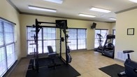 Located at 200-A Heron Cove Dr in Enterprise, Alabama, this single-floor fitness center offers a well-equipped and functional workout environment. The space is designed with practicality and performance in mind, providing residents with the essentials for a comprehensive exercise routine. Large windows with horizontal blinds line the walls, allowing abundant natural light to fill the room and creating an open, airy atmosphere. The light-toned tile flooring is complemented by rubber workout mats positioned beneath the equipment for added safety and stability. The strength training area features a robust power rack with an adjustable bench and a full barbell and weight plate setup, suitable for a wide range of resistance exercises. A wall-mounted television provides entertainment during workouts. On the cardio side, a stair climber or elliptical machine is available for low-impact aerobic training. The room's neutral, light-colored walls and bright recessed fluorescent lighting ensure a clean and energizing environment throughout the day. The overall layout is thoughtfully organized, maximizing the use of the available space while keeping the area uncluttered and accessible for all fitness levels.