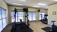 Located at 200-A Heron Cove Dr in Enterprise, Alabama, this single-floor fitness center offers a well-equipped and functional workout environment. The space is designed with practicality and performance in mind, providing residents with the essentials for a comprehensive exercise routine. Large windows with horizontal blinds line the walls, allowing abundant natural light to fill the room and creating an open, airy atmosphere. The light-toned tile flooring is complemented by rubber workout mats positioned beneath the equipment for added safety and stability. The strength training area features a robust power rack with an adjustable bench and a full barbell and weight plate setup, suitable for a wide range of resistance exercises. A wall-mounted television provides entertainment during workouts. On the cardio side, a stair climber or elliptical machine is available for low-impact aerobic training. The room's neutral, light-colored walls and bright recessed fluorescent lighting ensure a clean and energizing environment throughout the day. The overall layout is thoughtfully organized, maximizing the use of the available space while keeping the area uncluttered and accessible for all fitness levels.