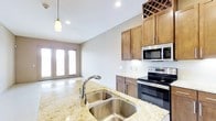 This single-floor space offers a thoughtfully designed open layout that seamlessly connects the kitchen and living areas. The kitchen is a standout feature, showcasing warm medium-toned wood cabinetry accented with sleek hardware, complemented by light granite countertops with subtle veining. A double-basin stainless steel sink sits at the island, providing both functionality and a central focal point. Stainless steel appliances, including a built-in microwave and electric range, add a polished, contemporary touch. A decorative wine rack integrated into the upper cabinetry adds a refined detail. Pendant lighting above the island and recessed ceiling lights throughout ensure the space is well-lit at all times. White subway tile backsplash ties the kitchen aesthetic together with clean, classic lines. The adjoining living area benefits from abundant natural light streaming through a set of glass-paneled French doors, which open to the exterior and enhance the sense of openness. The neutral wall tones and polished concrete-style flooring create a cohesive, modern ambiance throughout. The overall design balances practicality with style, making it a highly functional and visually appealing space.