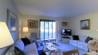 This single-floor space centers around a well-appointed living room designed for comfort and functionality. The room is furnished with a generously sized sofa and coordinating wingback chairs upholstered in classic floral fabric, arranged to encourage conversation and relaxation. A rectangular coffee table anchors the seating area, accompanied by an additional side table that adds practical surface space. Natural light streams generously into the room through large sliding glass doors, which open to an outdoor green area with mature trees visible beyond — a standout feature that connects the interior to the surrounding landscape. A wall-mounted air conditioning unit ensures year-round climate comfort. Entertainment is well accommodated with a flat-screen television positioned on a white media console along one wall. Subtle décor elements, including a framed artwork depicting two figures, add a personal touch to the neutral, light-toned walls. The flooring is carpeted throughout, contributing to the room's warm and relaxed atmosphere. Overall, the space presents a functional, lived-in character with practical amenities and a pleasant connection to the outdoors.