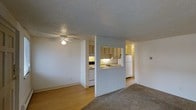 This single-floor residential space offers a practical and functional layout designed for everyday comfort. The living area is a welcoming space finished with plush carpeting and a ceiling fan with a light fixture, providing both comfort and ventilation. Adjacent to the living area, the flooring transitions to a smooth hardwood-style surface, creating a natural visual division between the relaxation and activity zones.

The kitchen is semi-enclosed and connected to the main living space via a convenient pass-through window, promoting an open and sociable atmosphere while maintaining a defined cooking area. The kitchen is equipped with white appliances, including a refrigerator and a range, along with ample upper cabinetry finished in a warm wood tone. The pass-through counter doubles as a casual bar or serving area, enhancing the kitchen's accessibility and utility.

The textured popcorn ceiling is consistent throughout the space, lending a classic residential character. Neutral wall tones and understated finishes give the space a clean, versatile aesthetic that allows for personalization. A hallway visible beyond the kitchen suggests additional rooms and storage, contributing to the overall functionality of this well-organized, single-level home.