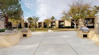 This residential community presents a thoughtfully landscaped outdoor environment that serves as a defining feature of the property. The grounds are characterized by expansive, well-maintained green lawns interspersed with mature pine and deciduous trees that provide generous shade throughout the common areas. Curved concrete walkways wind through the property, bordered by decorative river rock arrangements that add a naturalistic, Southwestern aesthetic to the landscape.

The buildings feature a classic stucco exterior in a neutral tan tone, complemented by Spanish-style clay tile rooflines that lend a cohesive architectural identity to the community. Balconies are visible on upper-level units, offering residents private outdoor spaces with views of the grounds.

A spacious concrete patio area serves as a communal gathering space, equipped with an outdoor BBQ grill station — ideal for outdoor dining and social activities. Nearby, a fenced pool area is visible through ornamental iron gates, framed by mature trees and shrub landscaping, providing a private and inviting recreational retreat.

The overall ambiance is relaxed and park-like, with the combination of verdant lawns, native-inspired rock borders, manicured shrubs, and open skies contributing to a comfortable and visually appealing outdoor living environment.