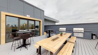 Located at 115 E 5th Ave in Columbus, Ohio, this rooftop amenity space combines bold interior design with a well-appointed outdoor deck. The interior lounge is immediately eye-catching, featuring a striking multicolored wood-slat ceiling in shades of yellow, gray, and natural tones that serves as the room's defining architectural element. Herringbone-patterned light wood flooring runs throughout, complementing the eclectic aesthetic. A built-in wet bar with sage-green cabinetry, a white quartz countertop, and a stainless steel sink lines one wall beneath a mounted flat-screen TV set against white subway tile. An exposed dark brick accent wall adds industrial character, while a cozy seating area with plush pink armchairs and a patterned area rug creates an inviting lounge nook. A glass-panel garage-style roll-up door opens to a window bar counter with wicker stools, offering views directly onto the outdoor deck. The expansive rooftop deck features a large wooden dining table with bench seating, multiple high-top tables and chairs, and dual stainless steel gas grills — ideal for gatherings and entertaining. The space is enclosed by sleek gray paneled walls, providing a modern, urban backdrop with open sky above.