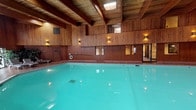This unique yoga studio is defined by its stunning indoor pool area, where thoughtful architectural design meets a warm, serene atmosphere. The pool itself is the centerpiece of the space, its vivid turquoise water shimmering beneath a dramatic ceiling of exposed wooden beams that rise to impressive heights. Natural light filters in through multiple skylights embedded in the ceiling, casting a bright, calming glow across the water's surface and enhancing the overall sense of openness and tranquility.

The walls throughout are clad in rich, warm-toned natural wood paneling that extends from floor to upper level, creating a cohesive and inviting aesthetic. Softly glowing wall sconces are mounted at regular intervals, providing a gentle ambient light that complements the natural illumination. A mezzanine-level overlook features additional windows that allow views into the pool area, adding depth and visual interest to the architecture.

The surrounding pool deck offers practical amenities, including lounge chairs for relaxation. A safety ring is mounted on the wall, and access doors connect to adjacent areas of the facility. The overall ambiance is one of calm, wellness, and natural warmth — an ideal environment for restorative practices, aquatic wellness, or mindful movement in a beautifully crafted, wood-accented setting.