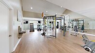 Located in Hoover, Alabama, this fitness center offers a comprehensive exercise facility designed to support a wide range of workout routines. The space is defined by its soaring vaulted ceilings, which lend an open, airy atmosphere, complemented by recessed lighting and large windows that invite abundant natural light and outdoor views of the surrounding landscape and pool area.

The flooring throughout consists of durable hardwood-style planks, providing a clean, modern aesthetic that is easy to maintain and suitable for diverse training activities. The layout is thoughtfully organized, with distinct zones for cardio, strength, and functional training.

The cardio section features multiple treadmills, an elliptical trainer, and a stair climber, each positioned in front of wall-mounted flat-screen televisions for an engaging workout experience. A dedicated functional training area houses a large multi-purpose training rig complete with TRX suspension straps, gymnastic rings, a heavy punching bag, and a cable pulley system. An assortment of kettlebells, resistance bands, and stability balls are neatly organized on an adjacent rack.

The strength training zone includes selectorized cable machines and weight benches, offering targeted muscle training options. Full-length mirrors line one wall, enhancing the sense of space while allowing users to monitor their form. The overall ambiance is bright, motivating, and professionally appointed, making this fitness center a standout amenity.