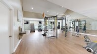 Located in Hoover, Alabama, this fitness center offers a comprehensive exercise facility designed to support a wide range of workout routines. The space is defined by its soaring vaulted ceilings, which lend an open, airy atmosphere, complemented by recessed lighting and large windows that invite abundant natural light and outdoor views of the surrounding landscape and pool area.

The flooring throughout consists of durable hardwood-style planks, providing a clean, modern aesthetic that is easy to maintain and suitable for diverse training activities. The layout is thoughtfully organized, with distinct zones for cardio, strength, and functional training.

The cardio section features multiple treadmills, an elliptical trainer, and a stair climber, each positioned in front of wall-mounted flat-screen televisions for an engaging workout experience. A dedicated functional training area houses a large multi-purpose training rig complete with TRX suspension straps, gymnastic rings, a heavy punching bag, and a cable pulley system. An assortment of kettlebells, resistance bands, and stability balls are neatly organized on an adjacent rack.

The strength training zone includes selectorized cable machines and weight benches, offering targeted muscle training options. Full-length mirrors line one wall, enhancing the sense of space while allowing users to monitor their form. The overall ambiance is bright, motivating, and professionally appointed, making this fitness center a standout amenity.