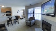 This single-floor residence features an open-concept layout that connects the living and dining areas, creating a cohesive and functional flow throughout the main living space. The living area is anchored by a fireplace with tile surround, positioned beside large windows fitted with horizontal blinds that allow ample natural light to fill the room. Neutral-toned carpet extends across the living and dining areas, contributing to a warm and understated ambiance. The dining area accommodates a dark wood table and chair set beneath a chandelier, complemented by a wall clock that adds a classic touch. Adjacent to the dining space is a breakfast bar with counter-height seating, offering a casual alternative for dining or entertaining. The open sightlines between each zone enhance the sense of space throughout the main level. Overall, the space presents a practical and comfortable layout with consistent neutral tones, natural light, and key functional features distributed across the connected living areas.