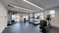 This fitness center offers a comprehensive workout environment designed to support a wide range of exercise routines. The space is finished with durable speckled rubber flooring throughout, providing both safety and functionality for various training activities. Sleek, recessed linear LED lighting runs along the ceiling, delivering bright, even illumination across the entire area.

The cardio zone is well-appointed with multiple treadmills and elliptical trainers from Life Fitness, as well as a recumbent bike, all positioned to take advantage of large windows that frame views of the surrounding neighborhood and fill the space with natural light. A multi-station cable machine anchors the strength training area, complemented by a full rack of dumbbells ranging across multiple weight increments and an adjustable workout bench.

Stability and functional training are supported by an assortment of exercise balls in varying sizes, resistance bands, and additional accessories neatly stored along the walls. A bold motivational typography mural — featuring phrases such as "Work Hard," "Will Power," and "Inspire" in black, white, and orange — adds an energizing visual focal point. Glass entry doors with branded signage contribute to the clean, modern aesthetic of this well-organized fitness facility.