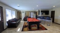 This single-floor space is designed with entertainment and socializing in mind. The open-concept layout connects the main living area seamlessly to a kitchen and bar, creating an inviting environment for gatherings. At the heart of the space sits a full-size pool table with a bold red felt surface, set atop a colorful area rug that anchors the room. Recessed lighting throughout provides consistent, even illumination across the tile flooring that spans the main area. The living space is furnished with dark leather sofas accented with decorative pillows, offering comfortable seating for guests. Wall décor, including a film reel accent piece, adds a playful, entertainment-themed character to the room. Large windows allow natural light to filter in, brightening the space. Adjacent to the living area, the kitchen features dark cabinetry, a built-in microwave, and a bar-height counter lined with dark bar stools, creating a natural gathering spot. The open sightlines between the kitchen and recreation area make the space feel expansive and well-connected. A hallway visible from the main room suggests additional rooms beyond, rounding out this well-appointed, recreation-focused layout.