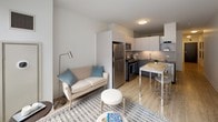 This well-appointed single-floor residence showcases a clean, contemporary design throughout. The open-concept layout seamlessly connects the living, dining, and kitchen areas, creating a cohesive and functional space bathed in natural light from large floor-to-ceiling windows that frame urban cityscape views.

The kitchen is outfitted with stainless steel appliances, including a gas range, over-the-range microwave, refrigerator, and dishwasher. Two-tone cabinetry — white uppers paired with grey lowers — complements the quartz countertops and a mosaic tile backsplash, while light wood-tone flooring ties the space together. A breakfast bar provides additional seating and a natural transition into the dining area.

The living area is bright and inviting, with light-toned flooring, neutral walls, and generous window exposure that brings in abundant daylight. The space comfortably accommodates a seating arrangement and media console, making it ideal for both relaxing and entertaining.

The bathroom is finished with a clean, modern aesthetic, featuring large-format tile flooring, a floating dark-toned vanity with an integrated sink, a wall-length mirror, and a glass-enclosed walk-in shower with white subway tile surround.

Throughout the unit, the consistent use of neutral tones, modern fixtures, and thoughtful design details creates a cohesive, contemporary living environment.