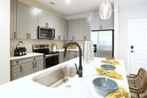 This single-floor residence at 3050 Circle 75 Pkwy SE in Atlanta, Georgia, presents a cohesive modern design throughout its well-planned layout. The kitchen is a standout feature, outfitted with gray shaker-style cabinetry, black hardware accents, light granite countertops, a neutral tile backsplash, and a full suite of stainless steel appliances. Wood-look plank flooring runs through the kitchen and extends into the living areas, adding warmth and continuity. The living space is generously proportioned, offering room for comfortable seating arrangements and benefiting from natural light through large windows dressed with layered curtains and blinds. The bedrooms are carpeted for comfort, each featuring windows with outdoor views and ample wall space. The primary bathroom is notably spacious, with gray cabinetry, tile flooring, and direct access to a walk-in closet equipped with wire shelving systems. A secondary bathroom mirrors the refined aesthetic, featuring a shower, vanity with gray cabinetry, and clean tile flooring. Throughout the residence, neutral tones are complemented by thoughtful design details, creating a polished and functional living environment.