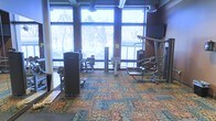 This fitness center offers a thoughtfully designed workout environment that balances functionality with an inviting atmosphere. The space is anchored by an impressive wall of floor-to-ceiling windows that flood the room with natural light and provide expansive views of the surrounding outdoor landscape, including mature trees — a refreshing backdrop for any workout session.

The facility is outfitted with a solid selection of strength-training equipment, including cable machines, leg press and extension stations, and a multi-function cable tower, catering to a range of exercise routines. The machines are arranged with ample space between them, allowing for comfortable movement and safe use. A wall-mounted flat-screen television adds a recreational element to the space.

Overhead, exposed industrial-style ductwork and ceiling beams contribute a modern, urban aesthetic, while mounted speakers suggest an audio system for an enhanced workout experience. The flooring features a distinctive, colorful patterned carpet with an organic, bubble-like design in earthy tones of blue, green, and orange — adding a lively visual element that sets this fitness space apart. The overall ambiance is bright, open, and welcoming, making it a practical and pleasant space for daily exercise.