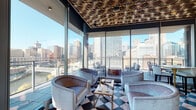 Located at 600 S Wells Street in Chicago's vibrant South Loop, this upscale lounge space delivers a striking blend of Art Deco elegance and contemporary design. The signature bold black-and-white checkered marble flooring anchors the entire space, creating a dramatic visual foundation that ties together the various seating areas.

Floor-to-ceiling glass curtain walls line the perimeter, flooding the interior with abundant natural light while offering sweeping, unobstructed panoramic views of the Chicago skyline, including the city's iconic towers and the Chicago River. A dedicated lounge zone near the glass walls features ornate, intricately patterned metallic ceiling tiles, adding warmth and visual depth overhead. Plush curved armchairs in muted tones are arranged around low cocktail tables, creating an inviting atmosphere for social gatherings.

The interior bar area is a focal point, featuring a sculptural curved service counter with warm wood cabinetry, glass-front storage, and pendant globe lighting suspended from coffered ceilings accented with warm wood beams. Additional seating clusters with leather swivel chairs and banquette-style sofas offer varied settings for guests. Curated artwork and decorative accessories punctuate the space, contributing to its refined, hospitality-forward ambiance. An adjacent balcony provides direct outdoor access with additional skyline views.