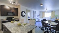 This single-floor space showcases an open-concept layout that integrates the kitchen, dining, and living areas into one cohesive, flowing environment. The kitchen is anchored by a generous granite-topped island with dark cabinetry overhead, offering both prep space and a natural dividing point between the cooking and living zones. A decorative round mirror adds a stylish focal point to the kitchen wall. The dining area sits adjacent, furnished with a dark table and modern seating. The living room extends comfortably beyond, featuring a ceiling fan, grommet-panel window treatments allowing natural light to filter in, and a curated arrangement of seating in white and green tones. Wall art and decorative accents contribute to the polished, contemporary ambiance throughout. A hallway visible from the main living area leads to a bedroom, hinting at a well-organized private zone separated from the shared living spaces. The consistent use of neutral white walls, dark flooring, and bold accent pieces creates a cohesive modern aesthetic that feels both inviting and well-appointed throughout the entire space.