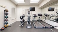 Located at Bristol Lakes in Mount Dora, Florida, this fitness center is a well-appointed, single-room exercise facility designed to support a variety of workout routines. The space features durable rubber flooring throughout, providing both safety and comfort during exercise. Natural light filters in through multiple windows fitted with blinds, as well as a skylight and a glass-paneled entry door, creating a bright and welcoming atmosphere. Large wall-mounted mirrors along one side of the room visually expand the space and allow users to monitor their form during workouts. The cardio section is equipped with treadmills, an elliptical trainer, and a stationary bike, complemented by a wall-mounted flat-screen television for entertainment. The strength training area includes an adjustable workout bench, a rack of dumbbells in graduated weights, and a set of medicine balls on a dedicated rack. Overhead fluorescent lighting supplements the natural light, ensuring consistent illumination throughout. The entry features a classic white paneled door with sidelights, and an additional interior door provides further access. The room's neutral beige walls and clean layout contribute to a focused, functional fitness environment suited for daily use by residents of the community.