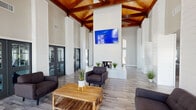 This single-floor community facility is defined by its striking architectural design and bright, open layout. The centerpiece of the space is a soaring great room with dramatic cathedral ceilings finished in warm, exposed wood beams and tongue-and-groove paneling, creating a rich contrast against the crisp white shiplap walls and tall structural columns. The great room features a wall-mounted flat-screen display above a sleek built-in fireplace, and is furnished with comfortable dark upholstered seating arranged around a rustic wood coffee table, offering an inviting gathering atmosphere. Large windows and glass-paneled double doors flood the area with abundant natural light. Adjacent to the great room, a dedicated workspace area provides desktop computer workstations set against the soaring backdrop, ideal for productivity. A separate conference or meeting room features a large dark stone-top table with sliding glass doors that open to an outdoor patio, connecting the interior seamlessly to the outdoors. A fitness center, accessible through glass-paned double doors directly from the great room, is visible and well-equipped. Throughout, durable wood-look plank flooring ties all areas together, while the consistent white shiplap detailing lends the entire space a cohesive, modern farmhouse aesthetic.