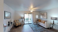 This single-story space centers around a well-appointed living room that combines comfort and functionality. The room features hardwood-style plank flooring that runs throughout, providing a clean and cohesive foundation. A ceiling fan with integrated lighting is centrally mounted, ensuring year-round comfort and ambient illumination.

Natural light is a standout feature, delivered generously through a full-width sliding glass door that opens to an outdoor patio area. The door is flanked by vertical blinds for adjustable light control, and the view beyond reveals lush greenery, creating a pleasant indoor-outdoor connection. A secondary window with vertical blinds adds further brightness to the space.

The living area is designed to accommodate a comfortable seating arrangement, with neutral-toned walls that complement a variety of décor styles. The textured ceiling adds subtle architectural character. Wall space is thoughtfully utilized, with room for artwork that enhances the overall aesthetic. The combination of natural light, open sightlines to the outdoors, and warm, neutral finishes gives the space an inviting and relaxed ambiance suitable for everyday living.