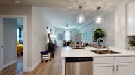 This single-floor residence offers a thoughtfully designed open-concept layout that seamlessly connects the kitchen, dining, and living areas. Light wood-look flooring flows throughout, creating a cohesive and contemporary aesthetic.

The kitchen is well-equipped with white shaker-style cabinetry, white quartz countertops, a classic subway tile backsplash, and a full suite of stainless steel appliances including a range, microwave, refrigerator, and dishwasher. A kitchen island with an undermount sink provides additional prep space and casual seating potential. Decorative pendant lighting adds a stylish touch overhead.

The open living and dining area benefits from natural light through multiple windows dressed with curtains and blinds. A ceiling fan anchors the living space, which offers comfortable room for both lounging and dining.

The bedroom is a relaxing retreat featuring large windows and a sliding glass door with vertical blinds, a ceiling fan, and wood-look flooring. The room offers generous space for furniture arrangement.

The bathroom maintains the home's clean, modern palette with white cabinetry, a wide vanity mirror, bright overhead lighting, and wood-look flooring, creating a fresh and functional space.