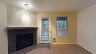 This two-floor space includes a living area that combines practical and comfortable design elements. The room features a wood-burning fireplace framed in dark tile, serving as a natural focal point along one wall. Carpeted flooring extends throughout the main area, adding warmth underfoot, while tile flooring flanks the fireplace surround. A glass-paneled exterior door floods the space with natural light and provides outdoor access, accompanied by a matching window fitted with horizontal blinds for privacy and light control. A wall-mounted heater unit is conveniently positioned beneath the window, offering supplemental climate control. The walls are finished in a neutral warm beige tone, complementing the textured popcorn ceiling. A track lighting fixture provides overhead illumination. The overall ambiance is understated and functional, with a neutral palette that offers a versatile backdrop for a variety of interior styles. The combination of the fireplace, exterior access, and ample window presence gives the living area a connected and inviting feel.