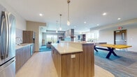 This thoughtfully designed single-floor amenity space seamlessly blends social, recreational, and functional areas into one cohesive, open-concept layout. The centerpiece of the kitchen area is a generous island topped with a crisp white quartz countertop and warm walnut cabinetry, illuminated by sleek pendant lights. The full kitchen includes stainless steel appliances — a French door refrigerator, dishwasher, and double sink — all complemented by a striking herringbone tile backsplash. Bar-height seating with modern navy and gold stools lines the island, creating an inviting gathering spot. Adjacent to the kitchen, a spacious lounge area is anchored by a dramatic floor-to-ceiling stacked stone fireplace with a linear gas insert and wood mantle, serving as a visual divider between two distinct seating zones. Plush sofas, accent chairs, and layered area rugs provide a warm, comfortable atmosphere, while large windows on multiple walls flood the space with natural light and offer expansive outdoor views. On the opposite end, a recreational zone houses a ping-pong table and additional seating, making the space equally suited for casual play or social events. Light hardwood flooring in the kitchen transitions to patterned carpet throughout the lounge and game areas, tying the varied zones together with a polished, contemporary aesthetic.