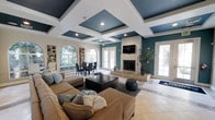 This single-floor space makes an immediate impression with its bold architectural design, most notably the coffered ceiling finished in a rich teal and crisp white color scheme that draws the eye upward and adds a sense of depth and elegance to the interior. Recessed lighting embedded within the coffers provides warm, even illumination throughout the space.

The open-concept layout seamlessly connects the living and dining areas, creating a spacious and cohesive environment. The living area is anchored by a large, comfortable sectional sofa arranged around a sleek, dark wood coffee table, all set atop a plush area rug over neutral tile flooring. A wall-mounted flat-screen television is positioned above a clean, white-framed fireplace with a raised stone hearth, serving as a natural focal point.

Teal accent walls complement the ceiling palette, tying the design together with a cohesive and contemporary feel. The dining area is positioned near a graceful arched window and French doors, which invite generous amounts of natural light and offer views of the surrounding greenery. Additional French doors on the opposite side of the room further enhance airflow and connectivity to the outdoors. Tall potted plants add a touch of organic warmth, rounding out this well-appointed and visually dynamic space.