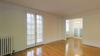 This single-floor space showcases a blend of classic architectural charm and bright, airy interiors. The main living area is anchored by gleaming hardwood floors that reflect abundant natural light streaming through a set of full-length French doors dressed with white plantation shutters. The doors lend an elegant, traditional character while offering excellent light control and privacy. Crisp white walls are framed by subtle molding details, adding a refined architectural touch throughout. An arched passageway connects the main living area to an adjacent room, providing a graceful transition that enhances the sense of flow and openness. The adjoining space is illuminated by its own window and features a vintage-style radiator, nodding to the building's period character. A delicate chandelier is visible through the archway, adding a decorative accent. The overall ambiance is clean, classic, and light-filled, with thoughtful architectural details — including panel molding, arched doorways, and period fixtures — that speak to the enduring quality of the space's original design.