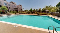 Located at 902 SE Brick Ave in Bentonville, Arkansas, this outdoor amenity space is centered around a generously sized rectangular swimming pool with clear turquoise water, framed by a clean concrete pool deck. The pool is equipped with stainless steel entry rails and safety life rings mounted along the perimeter fencing, ensuring a safe and functional environment. The surrounding deck offers ample space for relaxation, lined with sleek charcoal-toned chaise lounges paired with large shade umbrellas positioned on both sides of the pool. A dedicated lounge area on one end of the deck features a modular outdoor sofa set arranged around a wood-clad fire pit, creating an inviting social gathering spot. The space is enclosed by horizontal slat fencing that provides privacy while maintaining an open, airy feel. Lush plantings and mature trees soften the hardscape and add natural color throughout. The backdrop is defined by a striking multi-story building clad in warm wood paneling and dark metal siding, with brick accents contributing an urban-industrial character. The overall ambiance is modern and resort-inspired, offering a well-appointed outdoor retreat under the open Arkansas sky.