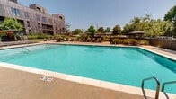 Located at 902 SE Brick Ave in Bentonville, Arkansas, this outdoor amenity space is centered around a generously sized rectangular swimming pool with clear turquoise water, framed by a clean concrete pool deck. The pool is equipped with stainless steel entry rails and safety life rings mounted along the perimeter fencing, ensuring a safe and functional environment. The surrounding deck offers ample space for relaxation, lined with sleek charcoal-toned chaise lounges paired with large shade umbrellas positioned on both sides of the pool. A dedicated lounge area on one end of the deck features a modular outdoor sofa set arranged around a wood-clad fire pit, creating an inviting social gathering spot. The space is enclosed by horizontal slat fencing that provides privacy while maintaining an open, airy feel. Lush plantings and mature trees soften the hardscape and add natural color throughout. The backdrop is defined by a striking multi-story building clad in warm wood paneling and dark metal siding, with brick accents contributing an urban-industrial character. The overall ambiance is modern and resort-inspired, offering a well-appointed outdoor retreat under the open Arkansas sky.