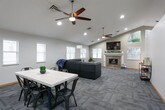 This single-floor space is thoughtfully designed to offer a warm and functional environment across several distinct areas. The entryway greets visitors with clean, neutral-toned walls, patterned carpet tile flooring, and light wood trim, establishing a professional yet welcoming atmosphere throughout.

The main lounge area is generously sized, featuring a large sectional sofa with colorful accent pillows and multiple windows fitted with white blinds that allow ample natural light to filter in. A comfortable seating nook off the main area includes a tile-surround fireplace with a white mantle, a wall-mounted television, and additional windows that enhance the bright, relaxed ambiance.

The kitchen is a standout feature of the space, boasting vaulted ceilings with track lighting, warm honey-toned wood cabinetry, granite-style countertops, and a peninsula with bar seating for two. Light wood-look flooring, a subway tile backsplash, and stainless steel appliances complete the functional and stylish design. The vaulted ceiling adds an airy, open quality that connects seamlessly to the adjoining common areas.

Throughout the space, the consistent use of neutral gray tones, natural wood accents, and patterned carpet tile creates a cohesive and inviting atmosphere well-suited for community gatherings or shared residential use.