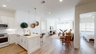 This single-floor residence is thoughtfully designed with a clean, modern aesthetic defined by crisp white walls, light wood-style flooring, and an abundance of natural light throughout. The open-concept layout seamlessly connects the kitchen, dining, and living areas into one cohesive, welcoming space.

The kitchen is a standout feature, outfitted with white shaker cabinetry, stainless steel appliances, a built-in wine cooler, and a peninsula island with a single-basin sink and brass fixtures. Gold pendant lights hang overhead, adding warmth to the bright, neutral palette. A built-in nook beside the kitchen provides additional display and storage space.

The living room receives generous natural light through large windows overlooking greenery, and is finished with a ceiling fan and warm hardwood-style flooring. The dining area connects directly to the kitchen, creating an ideal flow for everyday living and entertaining.

The bedrooms are bright and well-proportioned, each benefiting from large windows. The primary bathroom features a dual-sink vanity with dark cabinetry, a walk-in glass shower, and tile flooring. A spacious walk-in closet with built-in shelving and hanging rods provides ample storage. A secondary bathroom includes a vanity, tub-shower combination, and matching tile flooring.