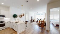 This single-floor residence is thoughtfully designed with a clean, modern aesthetic defined by crisp white walls, light wood-style flooring, and an abundance of natural light throughout. The open-concept layout seamlessly connects the kitchen, dining, and living areas into one cohesive, welcoming space.

The kitchen is a standout feature, outfitted with white shaker cabinetry, stainless steel appliances, a built-in wine cooler, and a peninsula island with a single-basin sink and brass fixtures. Gold pendant lights hang overhead, adding warmth to the bright, neutral palette. A built-in nook beside the kitchen provides additional display and storage space.

The living room receives generous natural light through large windows overlooking greenery, and is finished with a ceiling fan and warm hardwood-style flooring. The dining area connects directly to the kitchen, creating an ideal flow for everyday living and entertaining.

The bedrooms are bright and well-proportioned, each benefiting from large windows. The primary bathroom features a dual-sink vanity with dark cabinetry, a walk-in glass shower, and tile flooring. A spacious walk-in closet with built-in shelving and hanging rods provides ample storage. A secondary bathroom includes a vanity, tub-shower combination, and matching tile flooring.