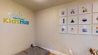 The Hawthorne Kids Hub is a thoughtfully designed, single-level children's activity space that provides a welcoming and stimulating environment for young visitors. The room features warm hardwood-style flooring and neutral, light-toned walls that create a bright and clean atmosphere conducive to play and creativity.

A standout feature of the space is a large gallery wall displaying a curated collection of modern geometric art prints in white frames, arranged in a bold grid pattern. The colorful, abstract designs add a vibrant and age-appropriate aesthetic to the room. Beneath the gallery, a child-sized round white table with matching chairs sits atop a patterned area rug, providing a dedicated space for arts and crafts, with art supplies readily available.

A wall-mounted wire rack organizer serves as a practical and accessible library, stocked with children's books, activity sets, and games. A cozy floor cushion offers a relaxed seating option for reading or lounging, and a woven basket nearby adds to the organized, tidy feel of the space.

The Kids Hub is directly adjacent to the fitness center, connected via a pass-through window and open doorway, allowing parents to supervise their children while exercising. The Hawthorne Kids Hub branding is prominently displayed on the walls, reinforcing the space's identity as a dedicated, community-focused children's area.