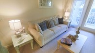 This single-floor space offers a welcoming and well-appointed interior centered around a comfortable living area. The living room is styled with a large, plush sofa upholstered in a classic ticking stripe fabric, generously adorned with an array of decorative pillows in coordinating neutral and blue tones, evoking a subtle coastal aesthetic. Soft, neutral wall tones create a warm and inviting ambiance throughout the space, while carpet flooring adds to the overall sense of comfort underfoot. Natural light filters gently into the room through a window dressed with sheer curtains, contributing to the bright and airy feel of the interior. A decorative mirror with a white geometric frame is mounted above the sofa, adding visual depth and a touch of elegance to the wall. The space is thoughtfully accessorized with side tables, ambient table lamps, and carefully chosen decorative accents, including what appears to be nature-inspired ornaments and a coffee table book. The overall ambiance is calm, cohesive, and designed for relaxed living, with attention to detail evident in the layered textures and harmonious color palette that tie the room together.