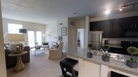 This single-floor residence is designed with an open-concept layout that seamlessly connects the kitchen and living areas, creating a cohesive and functional living space. The kitchen is equipped with dark espresso cabinetry that provides a striking contrast against the light granite countertops, which extend into a breakfast bar with seating. A stainless steel refrigerator and undermount sink contribute to the kitchen's modern aesthetic. The open counter design allows for clear sightlines into the living area, enhancing the sense of spaciousness throughout. The living area features neutral-toned carpeting and is furnished with comfortable seating arranged around a glass-top coffee table. Large windows along the far wall allow ample natural light to fill the room, brightening the soft, neutral wall tones. Framed artwork adds a decorative touch to the walls. A hallway visible from the main living space leads to additional rooms, suggesting a well-organized floor plan. Overall, the space presents a clean, contemporary ambiance with a warm and inviting atmosphere suited for everyday living.