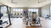 Located at 8801 Pathstone Blvd in Pensacola, Florida, this generously sized fitness center offers a well-equipped, single-level workout facility designed for a comprehensive exercise experience. The space features light wood-style flooring throughout, lending a clean and modern aesthetic that complements the neutral gray walls and crisp white trim.

Large, floor-to-ceiling and oversized grid-pane windows line the walls, flooding the interior with abundant natural light while providing views of the surrounding outdoor landscape, including palm trees and the neighboring community. Large ceiling fans are strategically positioned overhead to maintain airflow throughout the space, complemented by recessed lighting for consistent illumination.

The cardio section is well-appointed with a row of treadmills, ellipticals, rowing machines, and stationary bikes, all positioned to take advantage of the natural light and outdoor views. The strength training area features multiple multi-use cable machines, a Precor functional training system, weight benches, and a dumbbell rack. Full-length mirrors along one wall expand the sense of space and allow users to monitor their form during workouts.

The open layout ensures ample room to move freely between equipment stations, making the facility functional for simultaneous use by multiple individuals. A mounted flat-screen TV provides entertainment for cardio workouts, rounding out this thoughtfully designed fitness space.